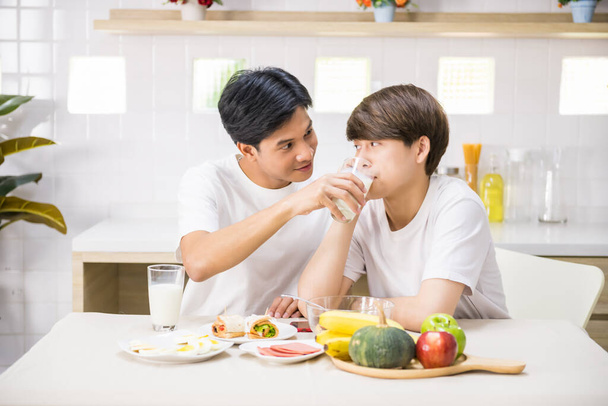 Der süße asiatische LGBT-Schwule füttert seinen Partner mit Milch, während er zu Hause Sandwich und Salat isst. Gleichgeschlechtliche Familie frühstückt. - Foto, Bild