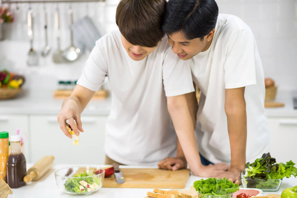 Joven pareja gay asiática hacer y cocinar ensalada de verduras juntos por dejar caer la manzana en un tazón en la cocina. Hombres LGBT encantadores juntos. Estilo de vida saludable para la familia del mismo sexo en San Valentín. - Foto, Imagen