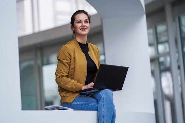 boldog mosolygós távoli dolgozó nő alkalmi ruhát ül keresztlábú pózol vele laptop vagy notebook az ölében egy modern szálloda előcsarnokában  - Fotó, kép