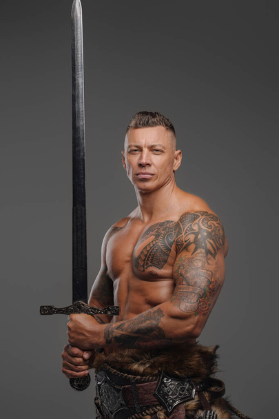Guerreiro muscular com torso nu segurando espada contra fundo cinza - Foto, Imagem
