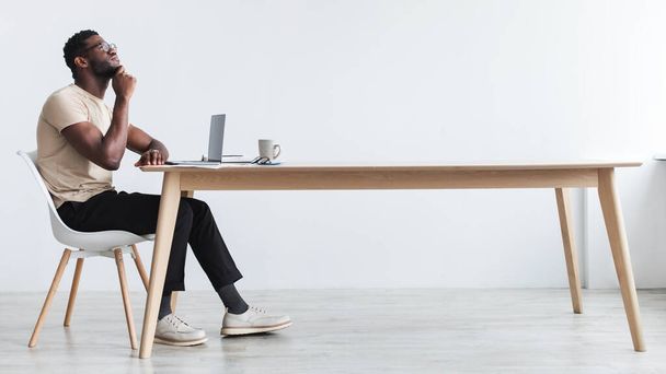 Pensive young black guy using laptop, sitting at desk, thinking over something, working online from home office - Фото, изображение