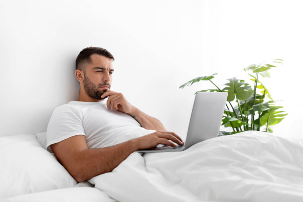Busy serious concentrated millennial muscular european man typing on computer, create idea, searching info, lies on bed - Photo, Image