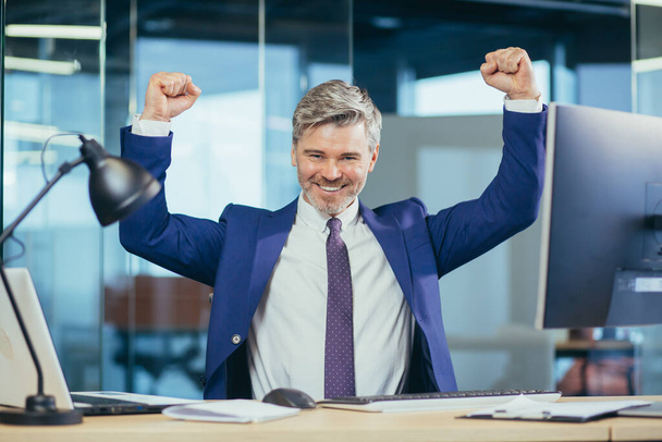 Successful boss works in a modern office at the computer, a senior man gray and happy with a beard looks at the camera and smiles, holds his hands up, celebrates victory - Fotografie, Obrázek