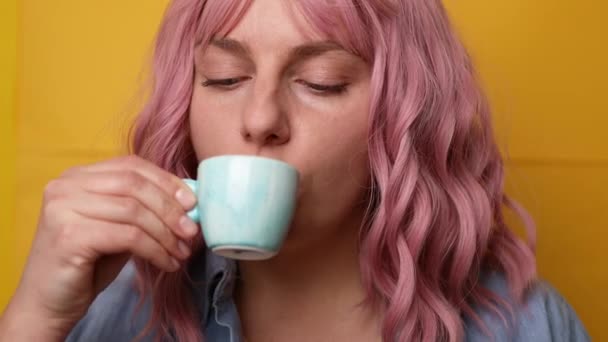 Portret radosnej młodej kobiety z różowymi włosami, cieszącej się filiżanką kawy na żółtym tle - Materiał filmowy, wideo