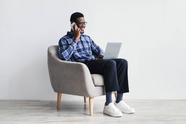 Χαρούμενος νεαρός Αφροαμερικανός με φορητό υπολογιστή που επικοινωνεί στο κινητό, κάθεται στην πολυθρόνα - Φωτογραφία, εικόνα