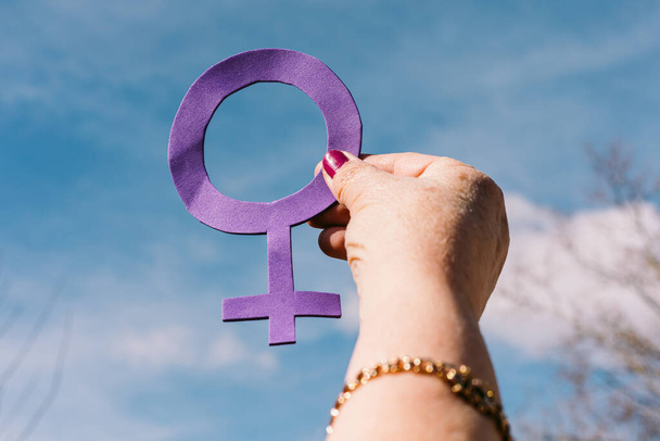 Hand of an older woman with purple nails, with the sky in the background, holding the female symbol in purple. Concept of women's day, empowerment, equality, inequality, activism and protest. - Photo, Image