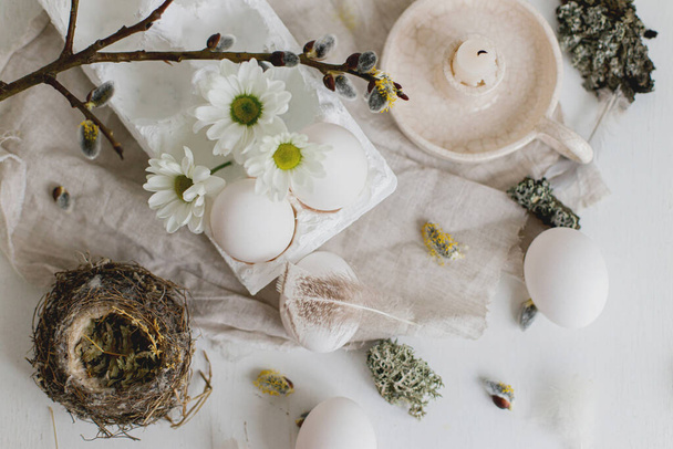 Natural easter eggs, feathers, willow branches, nest and candle on rustic white table flat lay. Stylish rural Easter still life in pastel white and beige colors. Modern simple aesthetics - 写真・画像