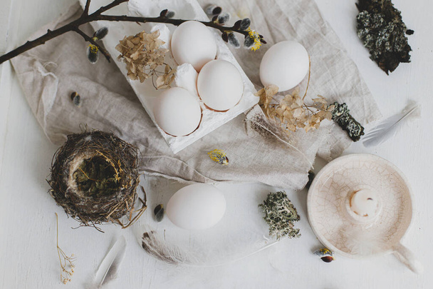 Pascua rústico piso laico. Huevos naturales, plumas, ramas de sauce, nido y vela en una mesa blanca envejecida. Elegante naturaleza muerta de Pascua rural en colores pastel blanco y beige. Estética simple moderna - Foto, imagen