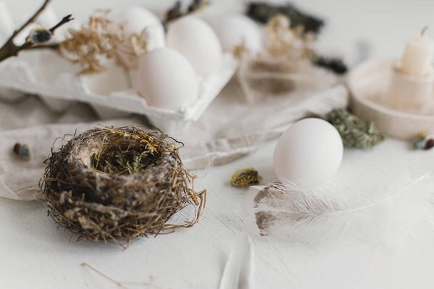 Easter simple aesthetics. Easter natural eggs, feathers, willow branches, nest on rustic white table. Stylish rural Easter still life in pastel white and beige colors. - Photo, Image