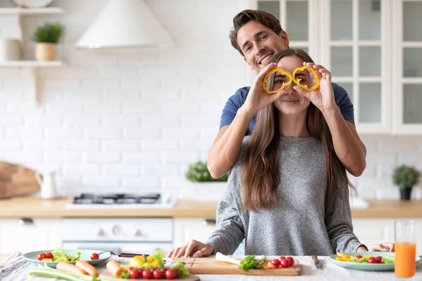 Positive young couple in love having fun while cooking in kitchen at home, making eyeglasses from bell pepper slices. Enjoying healthy preparing food together. - Foto, Imagen
