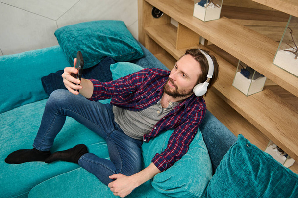 High angle view of handsome relaxed bearded Caucasian man wearing wireless headphones, talking by video call, looking at web camera on the smartphone in his hand, relaxing lying down on a sofa at home - Φωτογραφία, εικόνα
