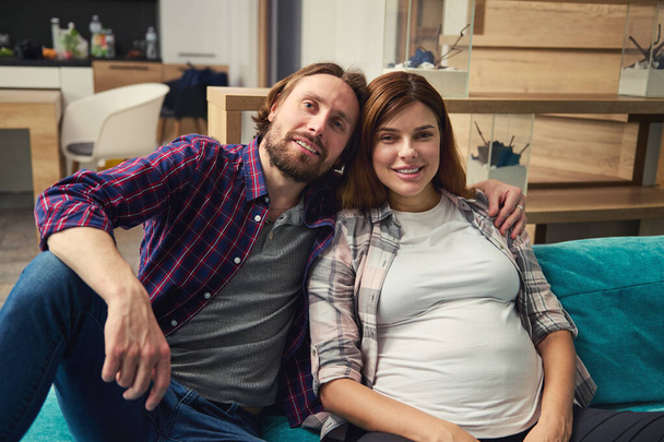 Портрет щасливої молодої кавказької пари в очікуванні, що дитина відпочиває разом на дивані вдома, посміхаючись, дивлячись на камеру. Прекрасна вагітна жінка, безтурботна вагітність. - Фото, зображення