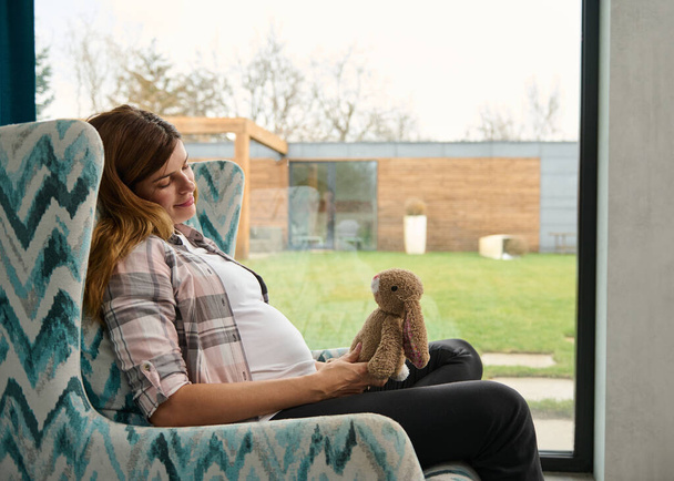 Serena mulher bonita esperando um bebê, sentado em uma poltrona e olhando sonhadamente para um brinquedo de coelho fofinho. Mulher grávida relaxando em casa contra janelas com vista para um jardim. Conceito de gravidez feliz - Foto, Imagem