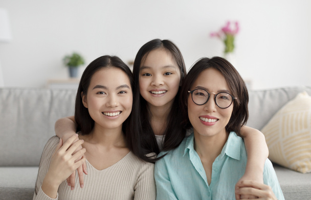 Portrait of joyful Asian family of mature mother, adult daughter and little granddaughter smiling at camera at home - Фото, зображення