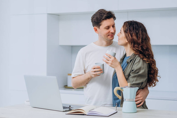 Married couple of freelancers took a coffee break in a bright kitchen standing at a table with a laptop - Фото, зображення