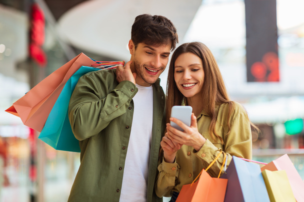 Coppia shopping utilizzando l'applicazione del telefono che tiene borse shopper nel centro commerciale - Foto, immagini
