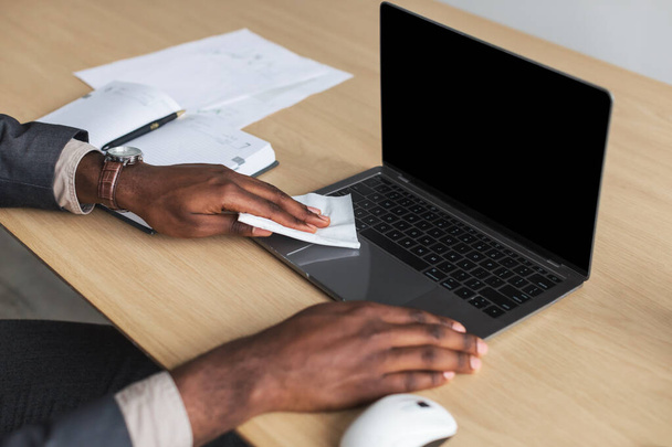 Неузнаваемый афроамериканский офисный работник, сидящий за столом, стирающий ноутбук салфеткой, макет для дизайна - Фото, изображение
