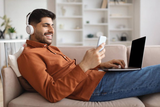 Uśmiechnięty mężczyzna w słuchawkach bezprzewodowych za pomocą telefonu komórkowego i komputera - Zdjęcie, obraz