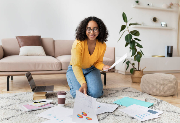 Прекрасна молода чорна жінка сидить на підлозі з ноутбуком і документами, працює або вчиться з дому, вільний простір
 - Фото, зображення