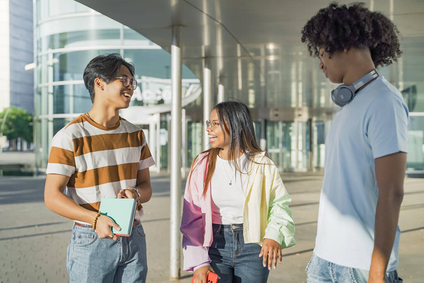 Junge multiethnische Freunde, die an einem sonnigen Tag glücklich auf dem modernen Universitätscampus spazieren und reden. - Foto, Bild