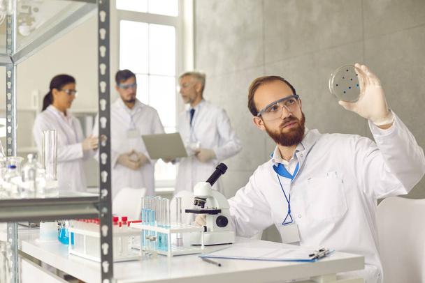 Male scientist analyzing test sample glass slide working at busy laboratory - Foto, Imagem