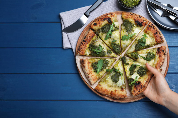 Woman taking slice of delicious pizza with pesto, cheese and arugula at blue wooden table, closeup. Space for text - Zdjęcie, obraz