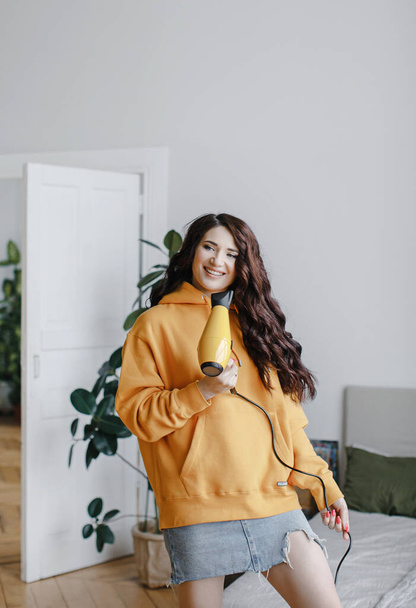 cute brunette girl in a yellow hoodie with a hair dryer poses, laughs for a beauty salon advertisement - Photo, Image