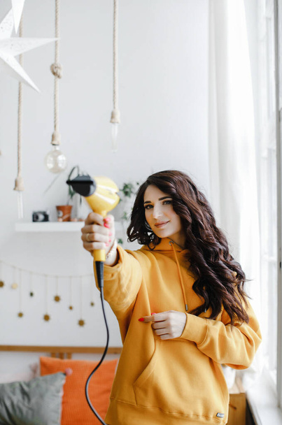 cute brunette girl in a yellow hoodie with a hair dryer poses, laughs for a beauty salon advertisement - Foto, immagini