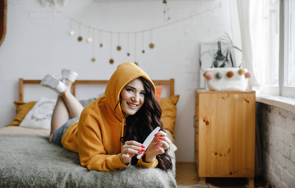 cute brunette girl in a yellow hoodie with a hair dryer poses, laughs for a beauty salon advertisement, pink manicure and nail file - Zdjęcie, obraz