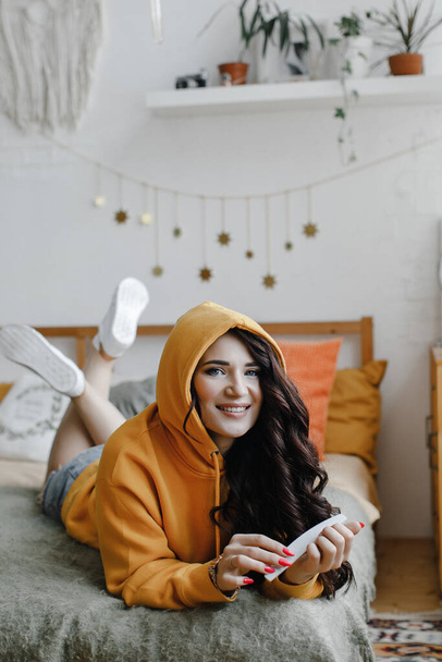 cute brunette girl in a yellow hoodie with a hair dryer poses, laughs for a beauty salon advertisement, pink manicure and nail file - Valokuva, kuva