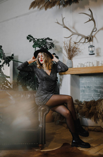 a beautiful sexy emotional girl in a short black shiny dress, hat and shoes poses in a stylish studio at a party - Photo, Image
