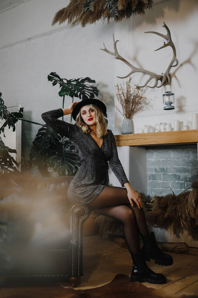 a beautiful sexy emotional girl in a short black shiny dress, hat and shoes poses in a stylish studio at a party - Фото, зображення