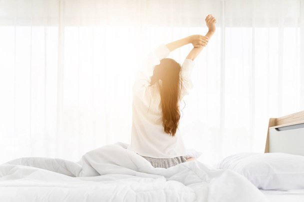 Jó reggelt, új nap. Ázsiai nő felébred, és ül test nyújtózkodik az ágyon ablak mellett a hálószobában - Fotó, kép