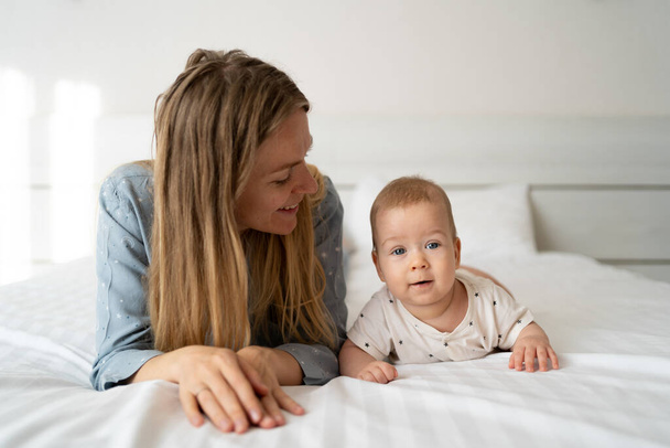 Portrait of a mother with a four month old baby in the bedroom. Young happy mum lies next to her adorable baby son, a cute blond boy playing in a white bed, enjoying a sunny morning in a home bedroom - Photo, Image