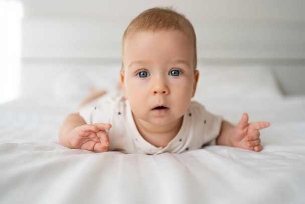 青い目をした赤ん坊の肖像画は、白いベッドの上に横たわり、ボディスーツを着て、 6ヶ月の赤ちゃん、寝室の白い背景の幼児を世話しています。赤ちゃん衛生の概念 - 写真・画像