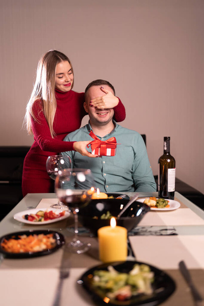 A young woman makes a gift to her lover and closes his eyes with her hand. A handsome man receives a surprise gift box from his girlfriend at dinner. Valentine's day at home. birthday gift concept - Photo, Image