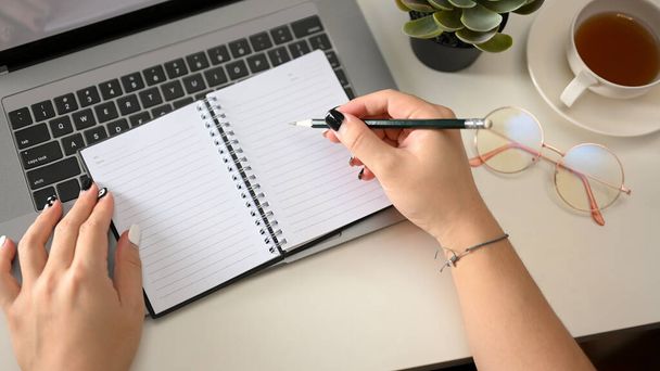 Γυναικεία χέρια κρατώντας στυλό και γράφοντας σε άδειες σελίδες σημειωματάριου στο μοντέρνο τραπέζι του γραφείου της. πάνω όψη - Φωτογραφία, εικόνα