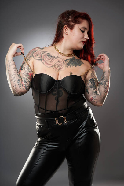 Glamour plus size model covered in tattoos in lingerie and leather on gray background - Valokuva, kuva
