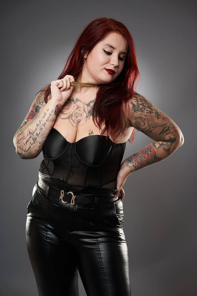 Glamour plus size model covered in tattoos in lingerie and leather on gray background - Foto, Imagen