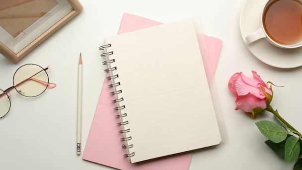 Όμορφη θηλυκό γραφείο θηλυκό γραφείο με άδειο κάλυμμα σημειωματάριο mockup, ροζ χαρτί, ροζ τριαντάφυλλο, φλιτζάνι τσάι, και διακόσμηση σε λευκό τραπέζι. πάνω όψη - Φωτογραφία, εικόνα