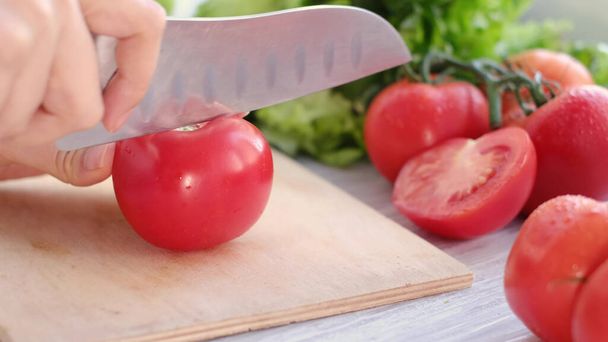 hands using kitchen knife cutting fresh tomato on wooden cutting board, with sprig of tomatoes and lettuce on background - Foto, Bild