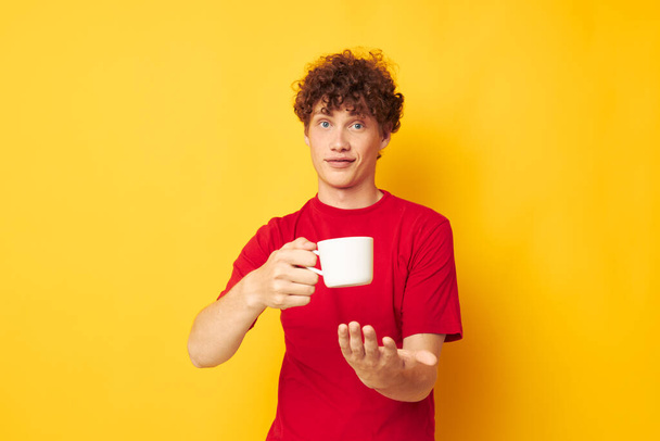 Young curly-haired man posing with a white mug and in the hands of a drink yellow background unaltered - Photo, image