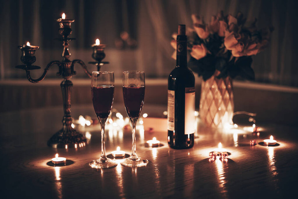 Een date bij kaarslicht. Bril met wijnkaarsen boeket van roos en geschenkdoos op tafel. Romantisch diner bij kaarslicht 's avonds thuis - Foto, afbeelding
