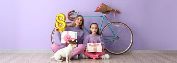Dziewczynka z matką, ładny pies, rower i prezenty w pobliżu ściany liliowej. Międzynarodowy Dzień Kobiet - Zdjęcie, obraz