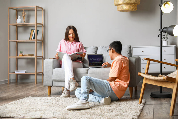 Молодой человек бронирует гостиничный номер онлайн, пока его жена читает журнал дома - Фото, изображение
