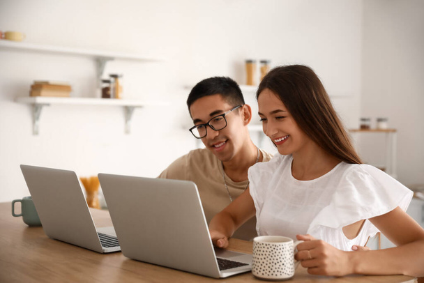 Giovane donna con tazza di caffè e suo marito utilizzando computer portatili a tavola in cucina - Foto, immagini