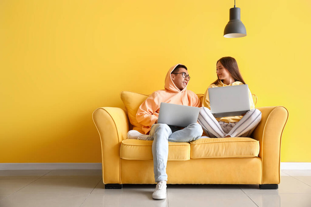 Νεαρό ζευγάρι με φορητούς υπολογιστές κάθεται σε καναπέ κοντά σε κίτρινο τοίχο - Φωτογραφία, εικόνα