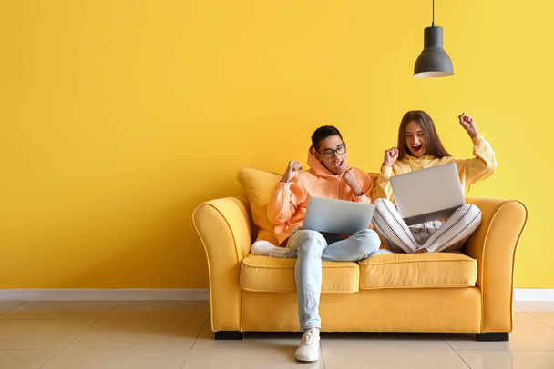 Ευτυχισμένο νεαρό ζευγάρι με φορητούς υπολογιστές που κάθονται σε καναπέ κοντά σε κίτρινο τοίχο - Φωτογραφία, εικόνα