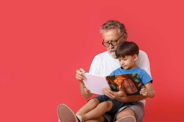 Vanhempi mies lukemassa kirjaa pienelle pojanpojalleen punaisella taustalla - Valokuva, kuva