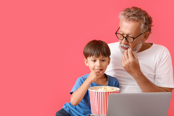 Küçük çocuk ve dedesi kırmızı arka planda video izlerken patlamış mısır yiyorlar. - Fotoğraf, Görsel